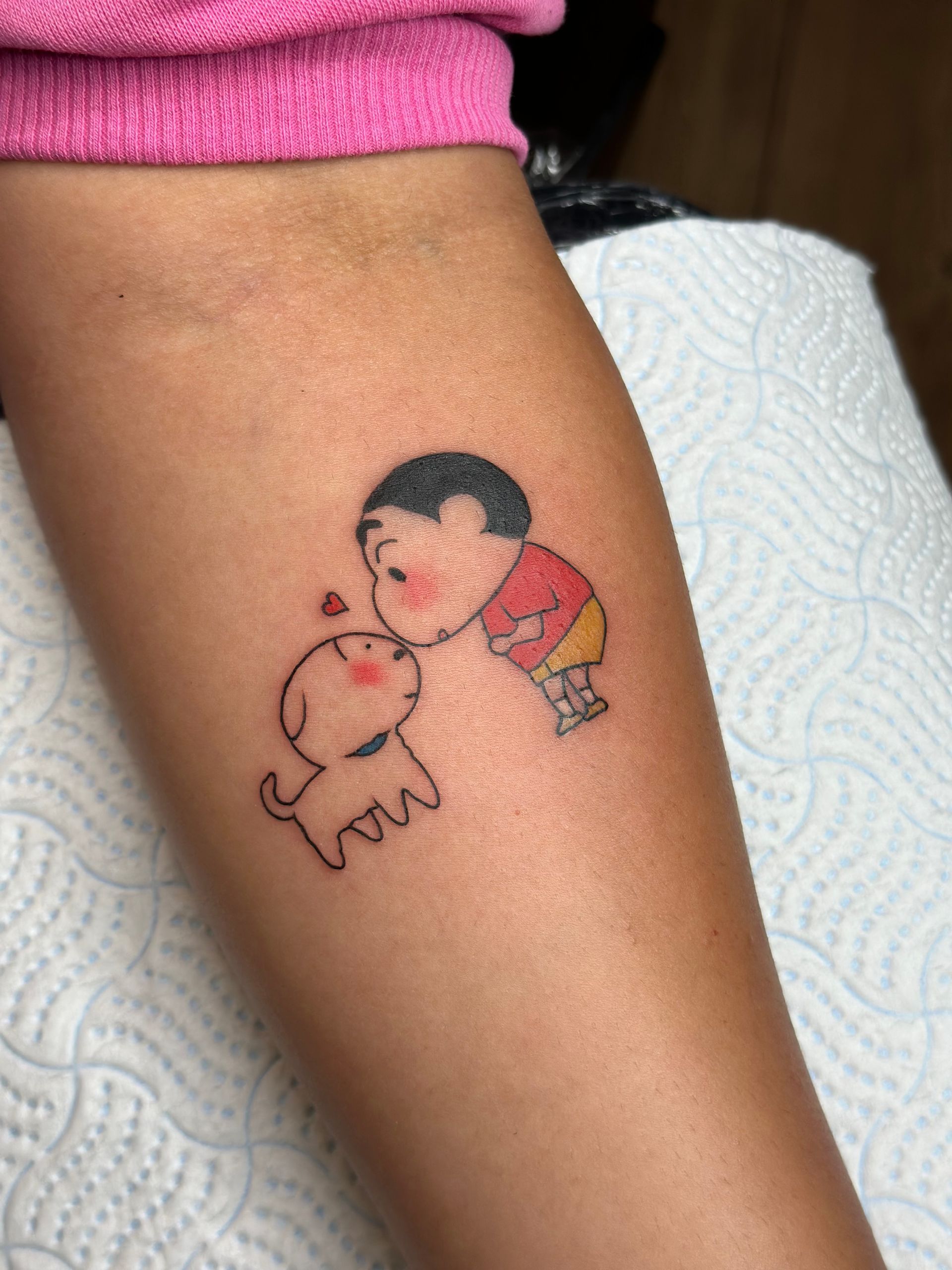 A colorful, small tattoo on an arm depicting the cartoon character Shin-chan leaning down to kiss his puppy, Shiro.