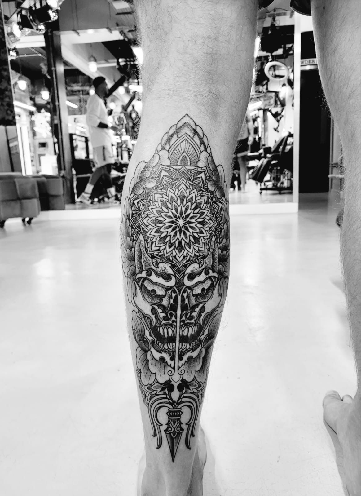 A detailed black-and-grey tattoo featuring a patterned mandala above a demonic skull mask on a person's lower leg.