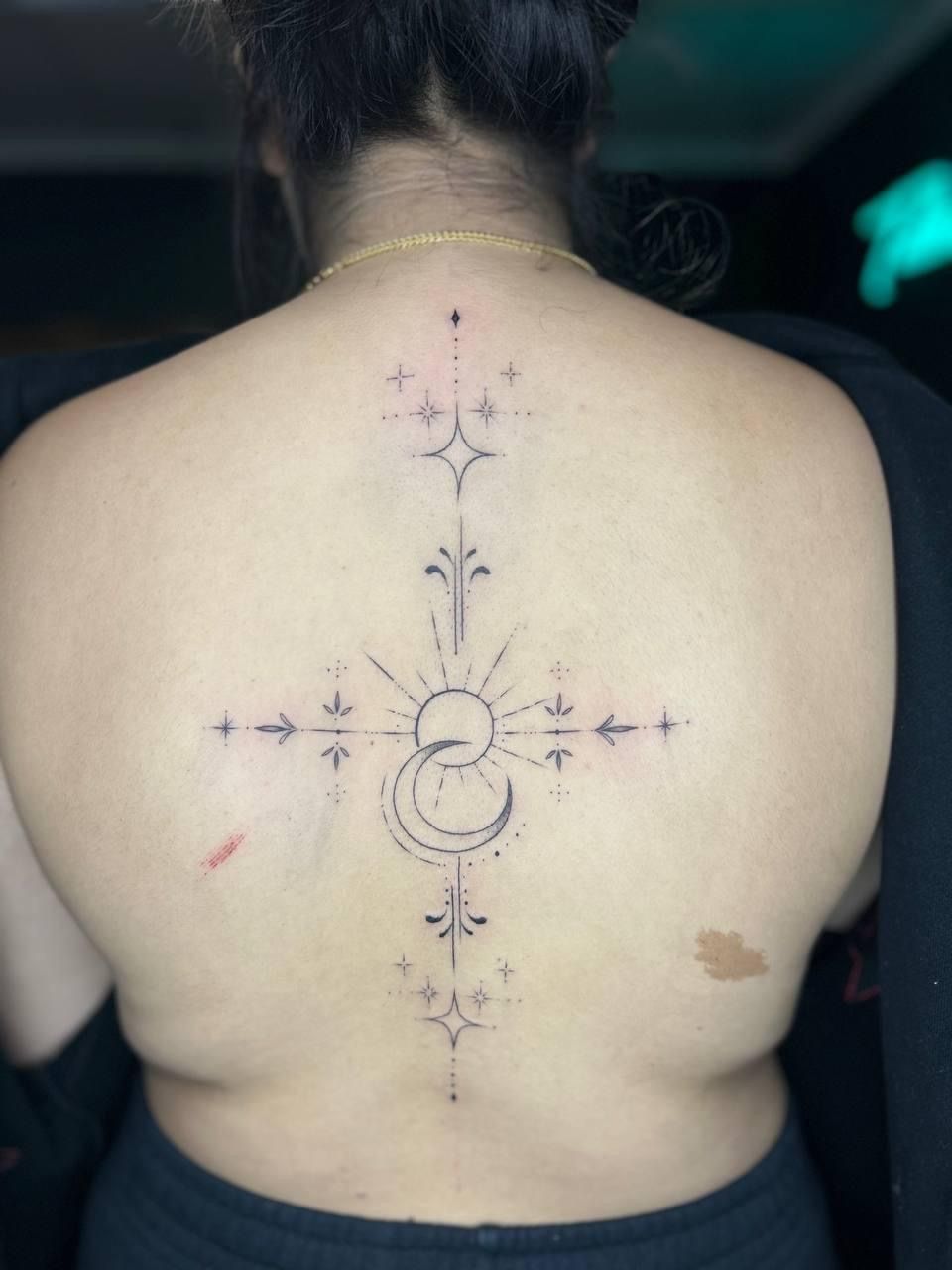 A fine-line back tattoo featuring a celestial design with a sun, crescent moon, and stars arranged in a vertical cross.