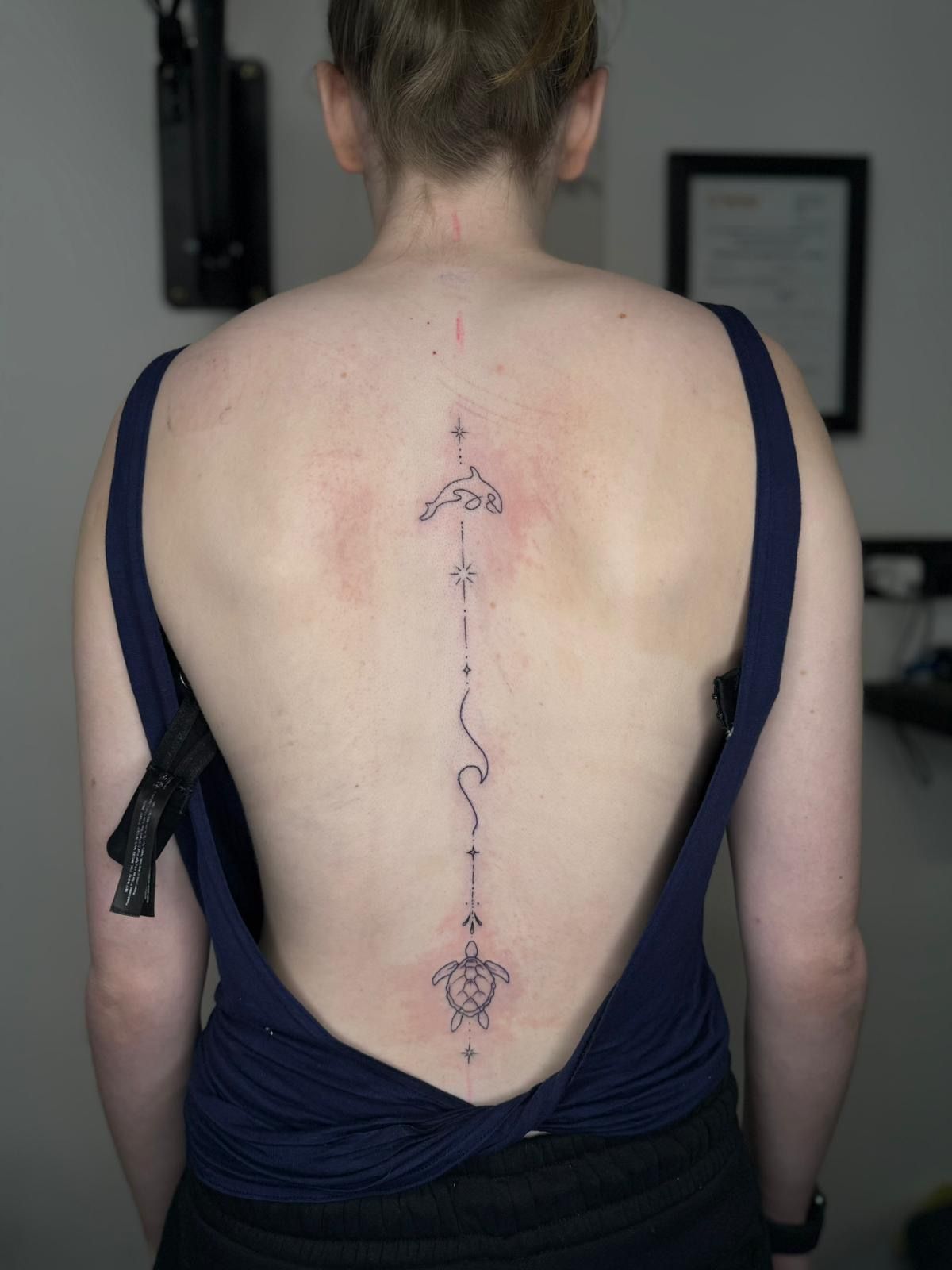 A person's bare back displaying a fine-line, vertical tattoo featuring a dolphin, wave, and turtle design.