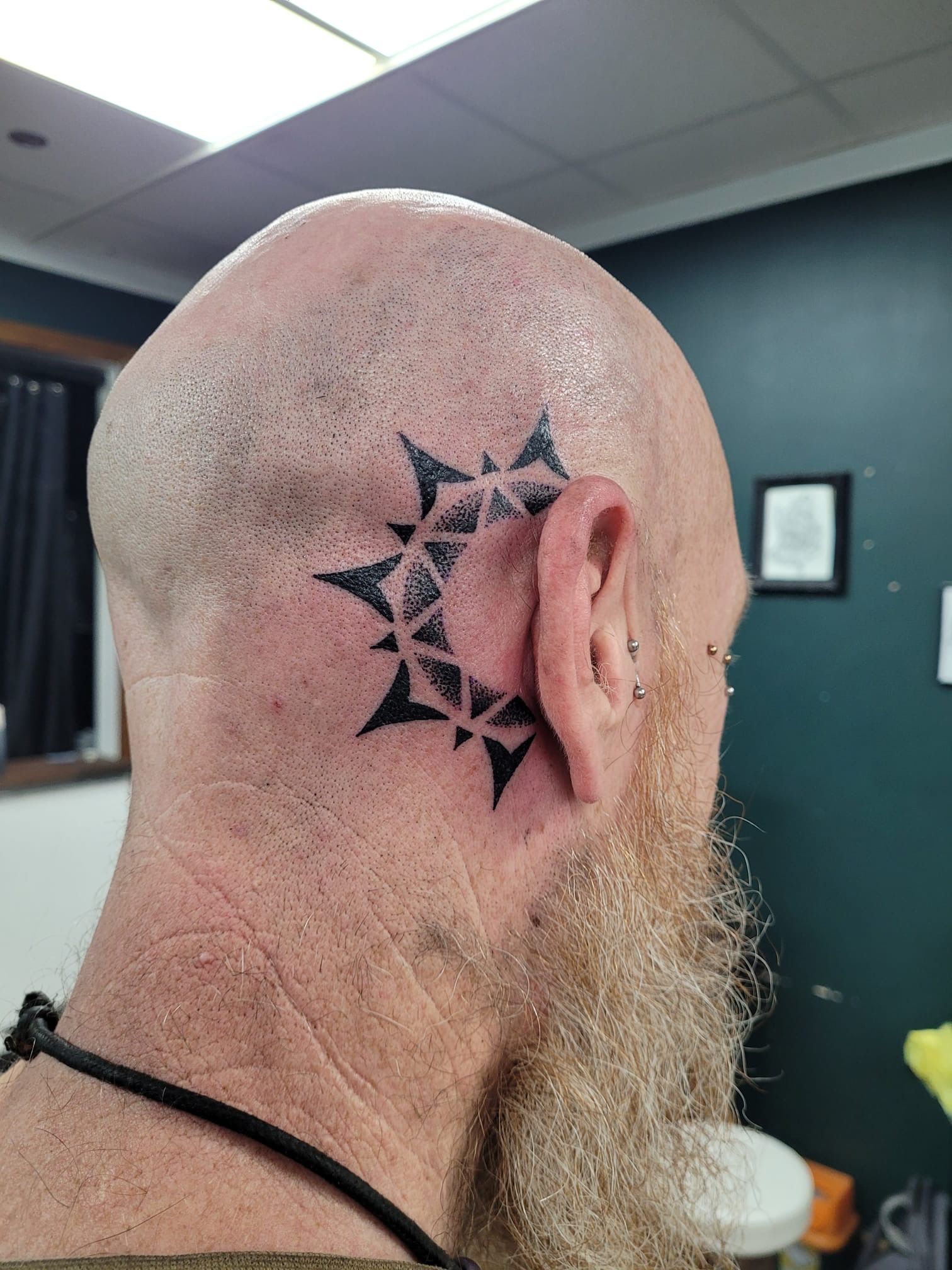 A close-up of a black geometric, star-shaped tattoo on the side of a bald head behind the ear.