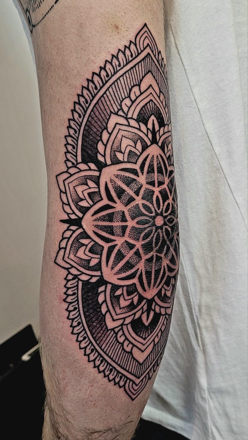 A black ink mandala tattoo wraps around the side of an elbow, featuring intricate geometric patterns and stippled shading.