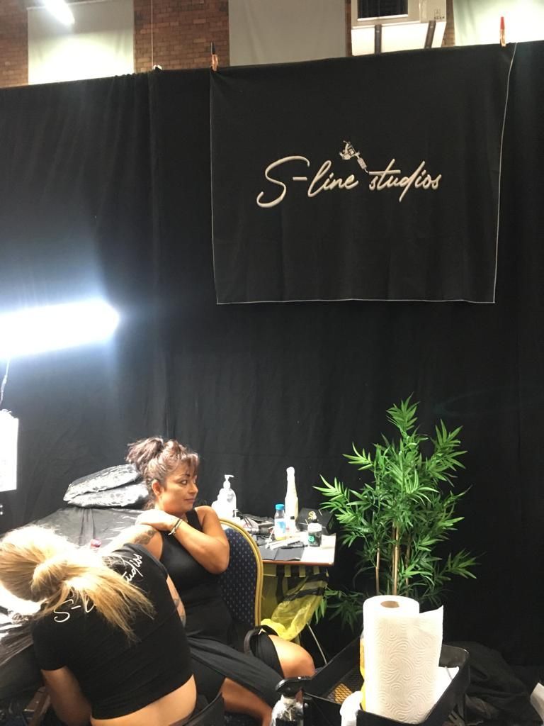 A tattoo artist works on a client’s arm in a booth with a black backdrop featuring an 