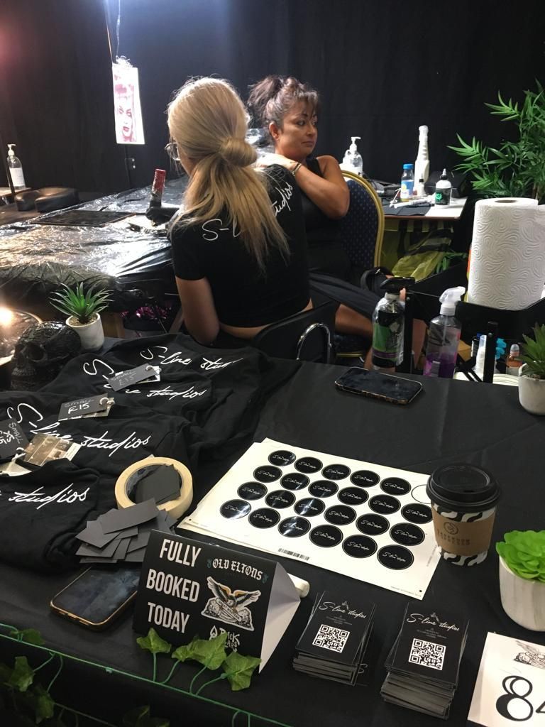 A tattoo artist works on a client in a studio filled with branded merchandise, stickers, and a 