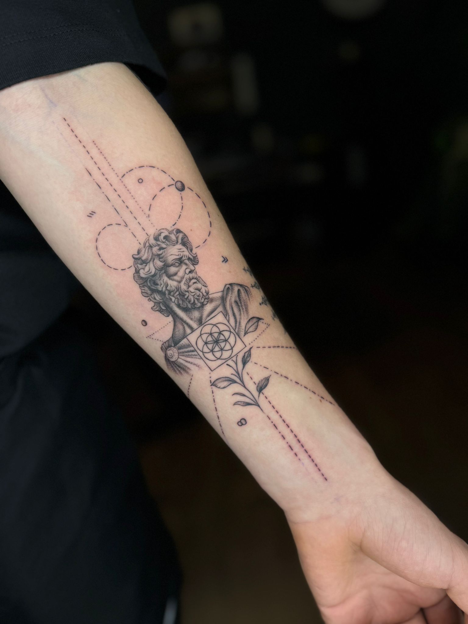 A detailed black ink forearm tattoo of a classical bust sculpture, surrounded by delicate geometric lines and dots.