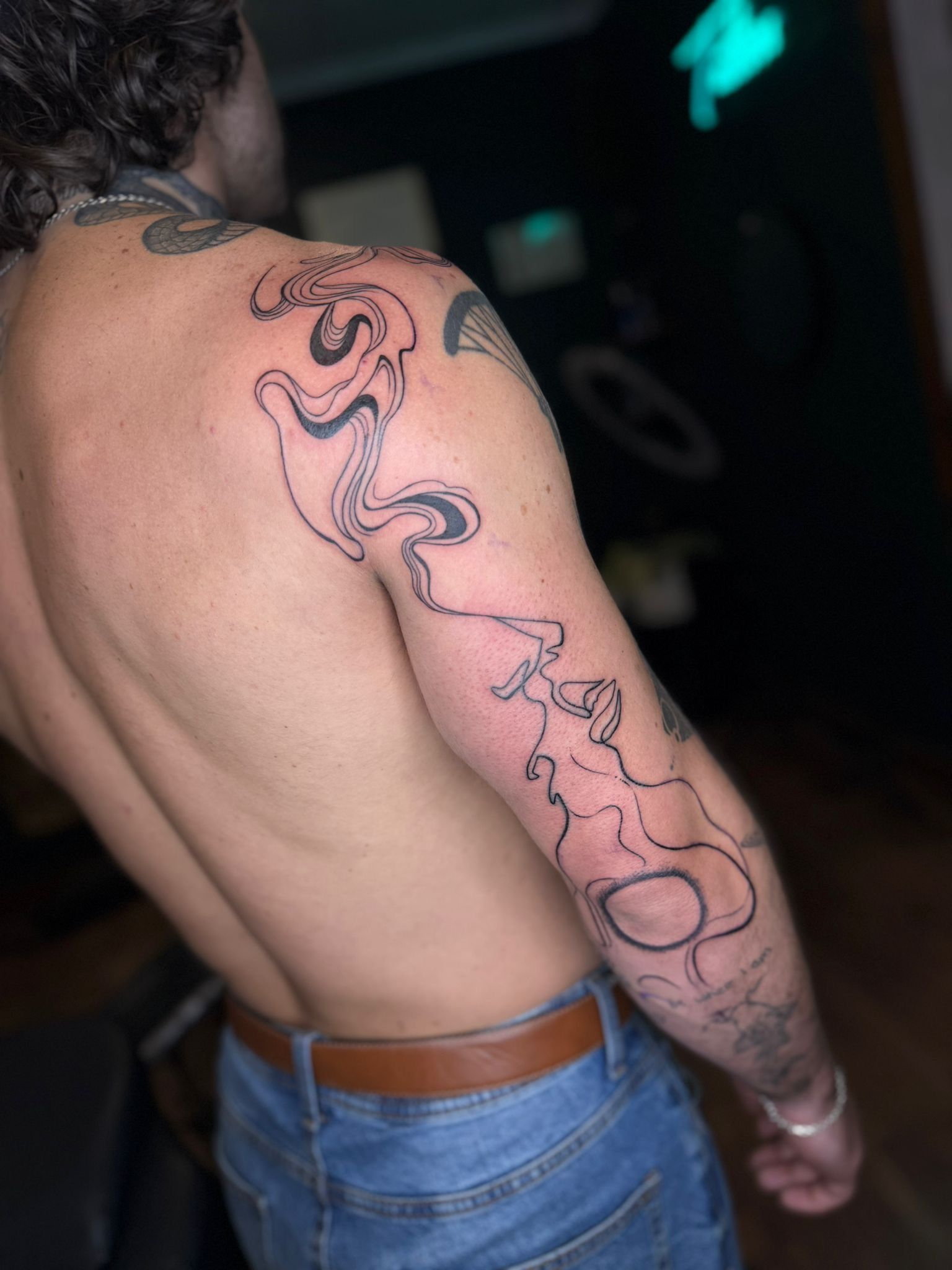 A tattoo on a person's upper arm featuring a stylized, free-flowing line drawing of an abstract figure.