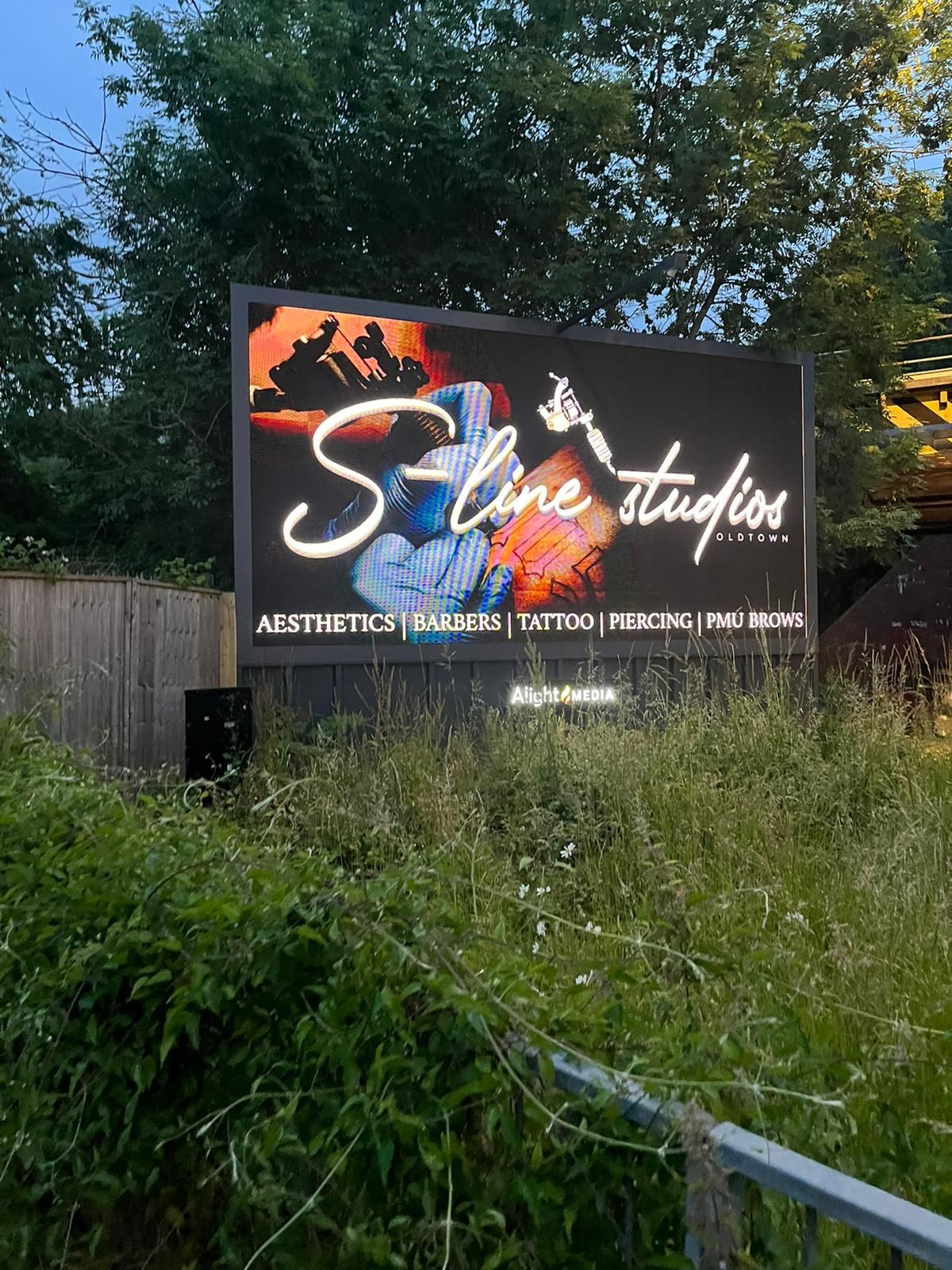 A neon-style sign for S-Line Studios, listing services including aesthetics, tattoo, and piercing, set in tall grass.