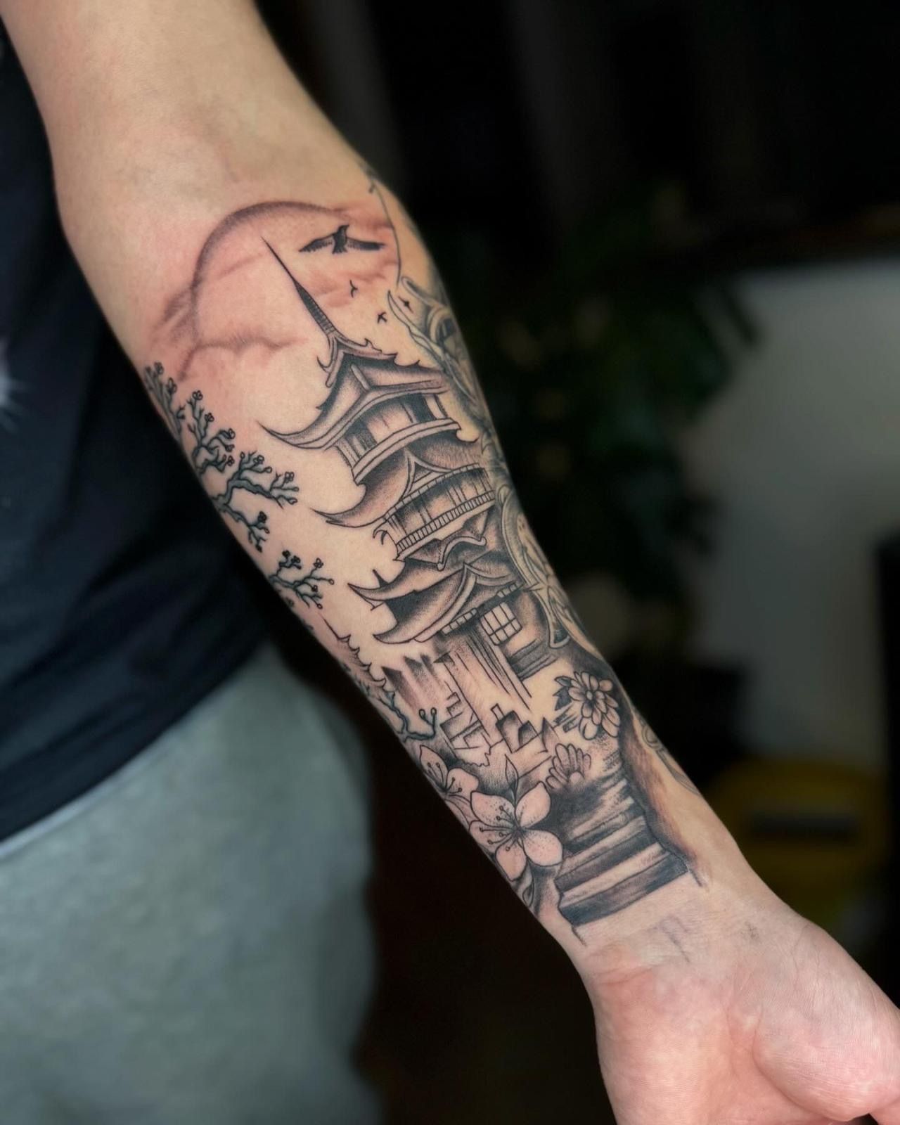 A detailed black-and-gray tattoo on a forearm featuring a Japanese pagoda, cherry blossoms, a rising sun, and birds.