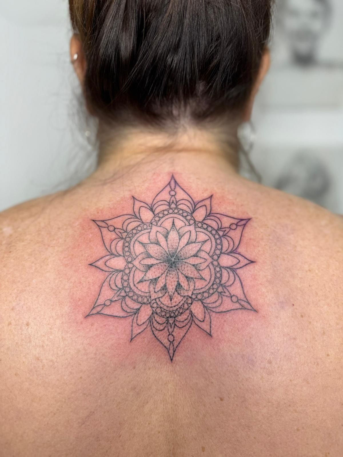 A black-ink, symmetrical mandala tattoo on the upper back, showing thin linework and delicate geometric details.
