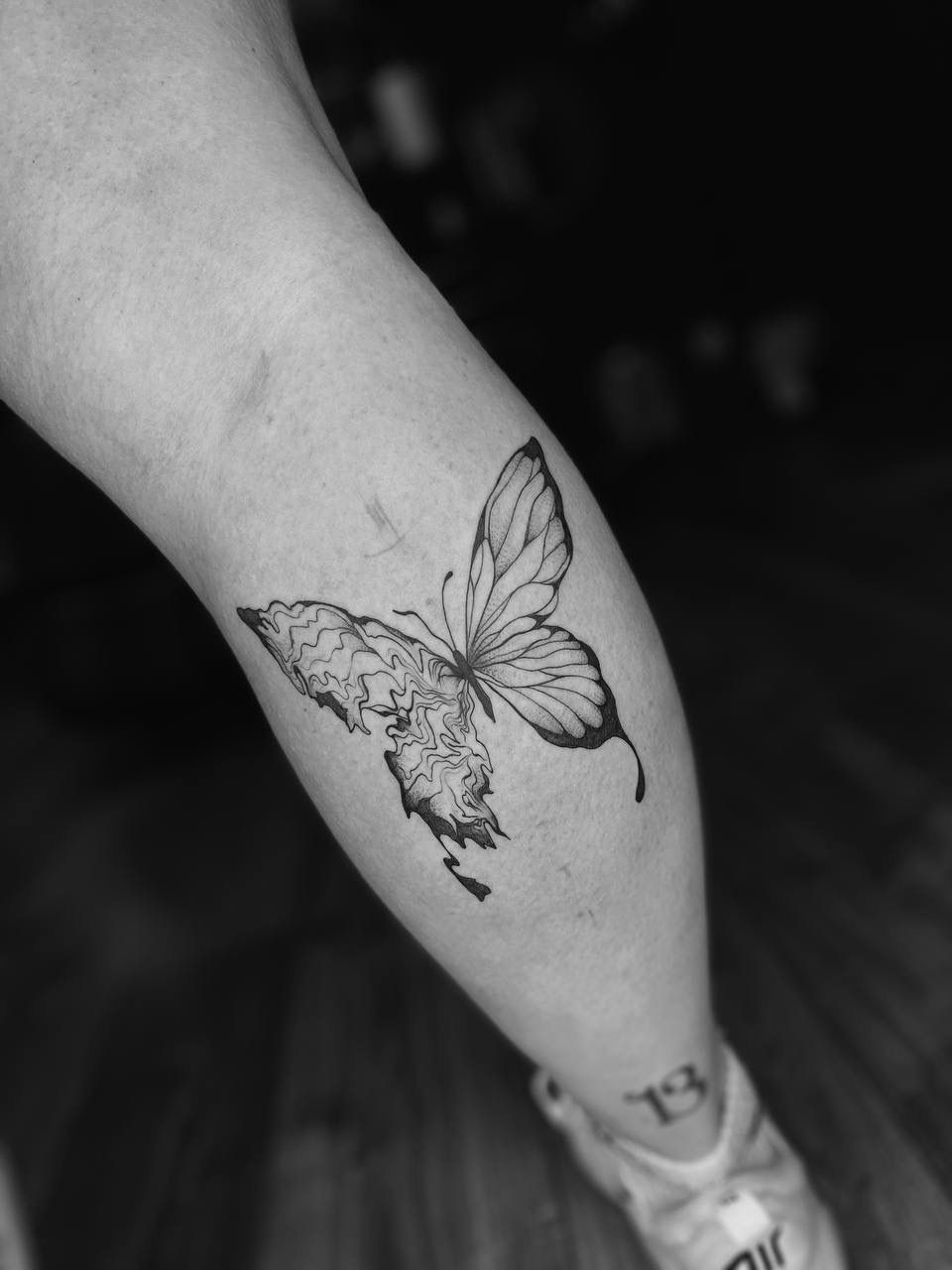 A black-and-white butterfly tattoo on a person's lower leg, featuring a detailed wing design on one side.