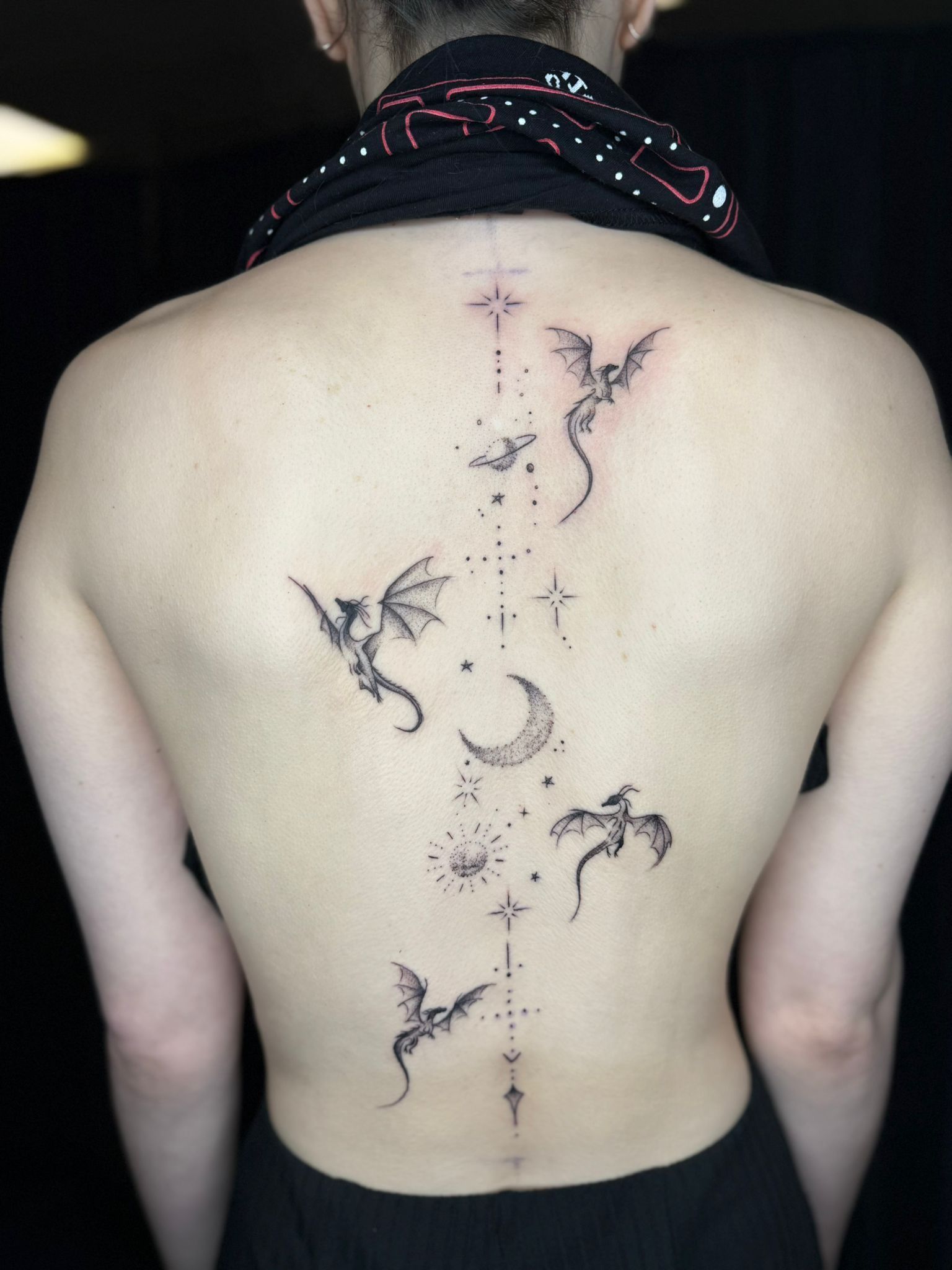 A person’s back with a delicate spine tattoo featuring multiple small dragons, stars, a crescent moon, and sun symbols.