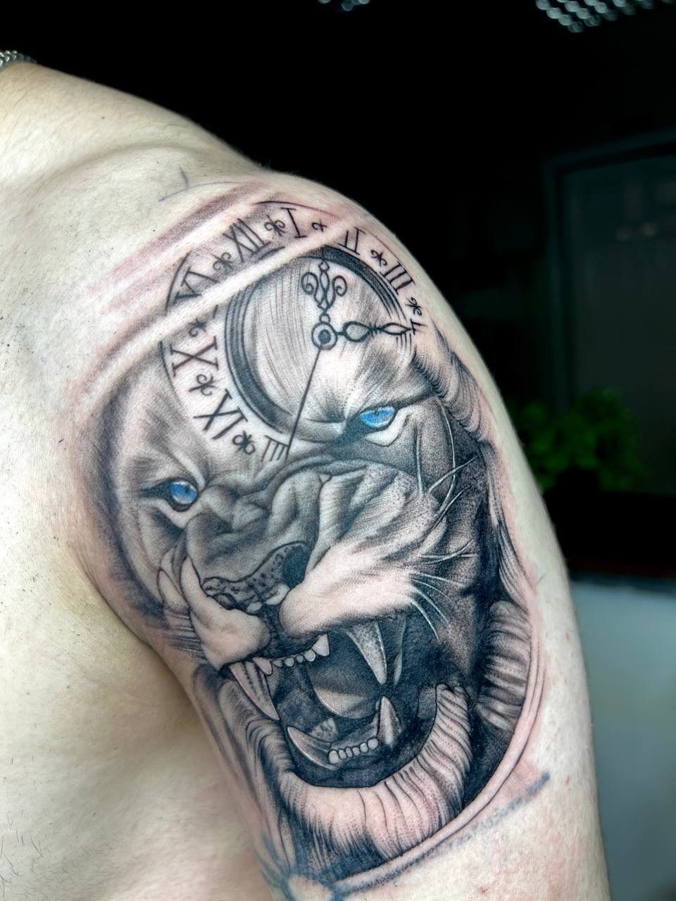A shoulder tattoo of a snarling lion with piercing blue eyes, overlaid with a clock face featuring Roman numerals.
