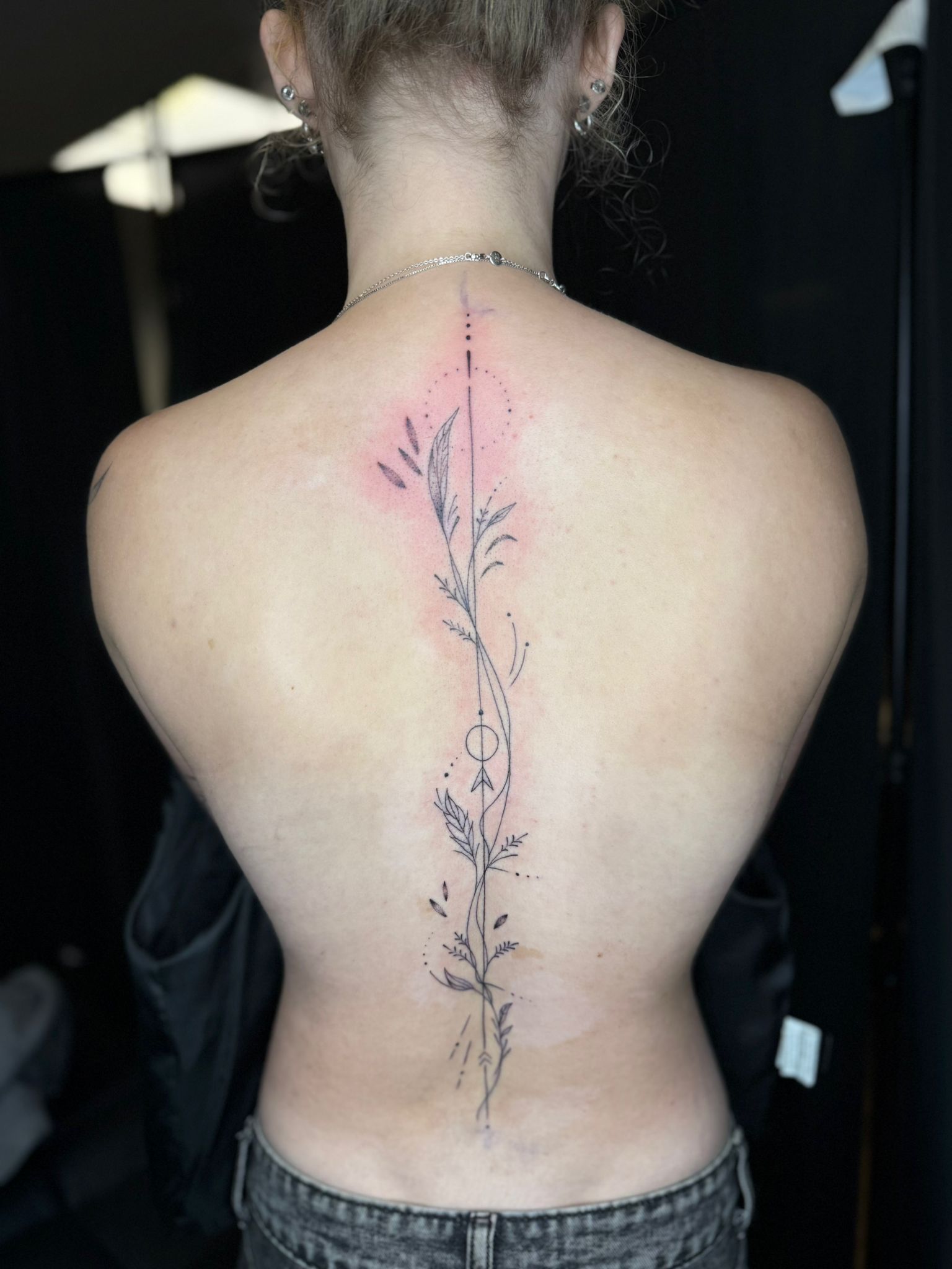 A person's back showing a vertical, fine-line tattoo along the spine featuring botanical sprigs and geometric accents.