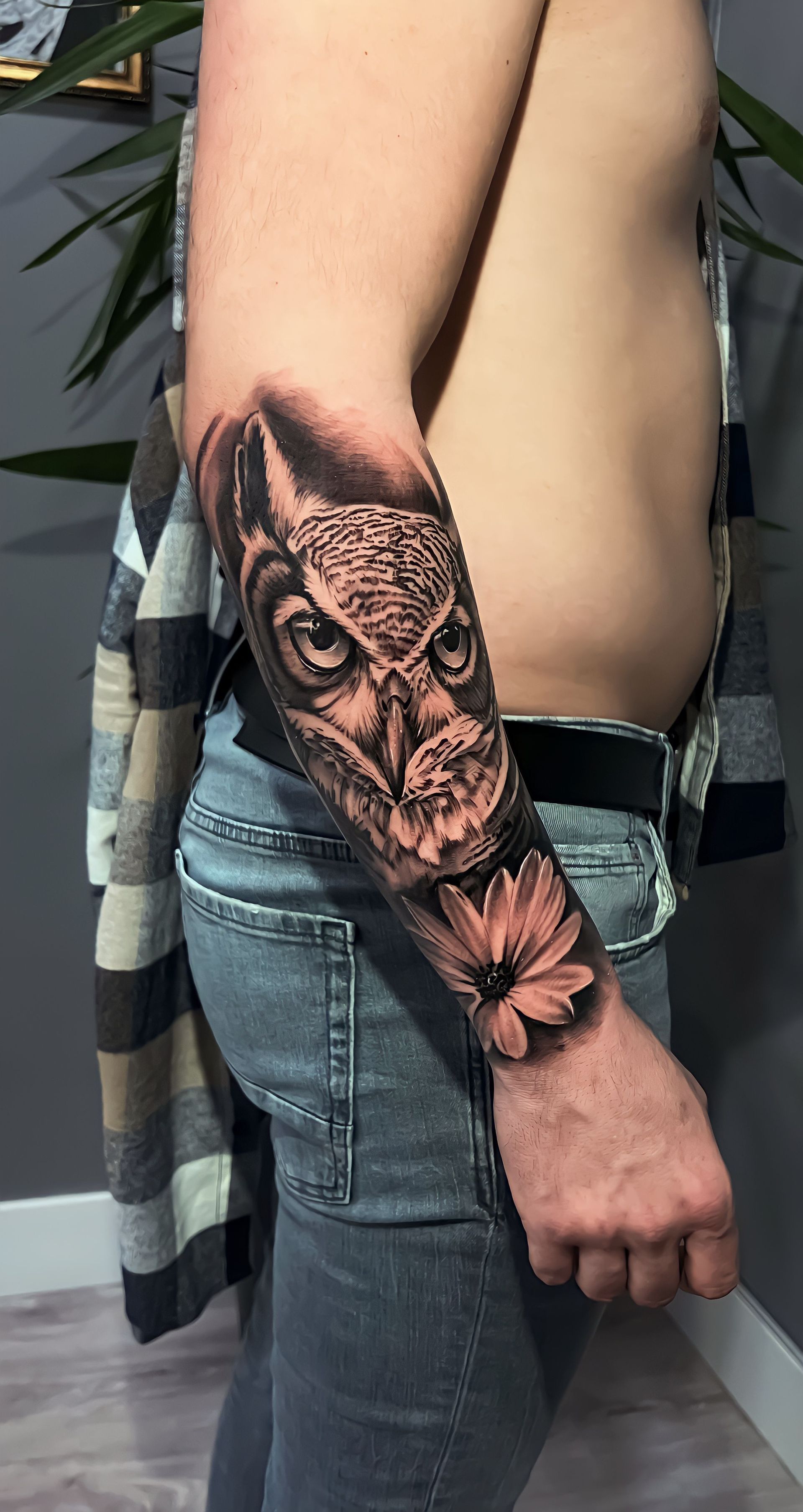 A tattoo of an owl's face paired with a flower on a person's forearm, shown in side profile against a dark background.