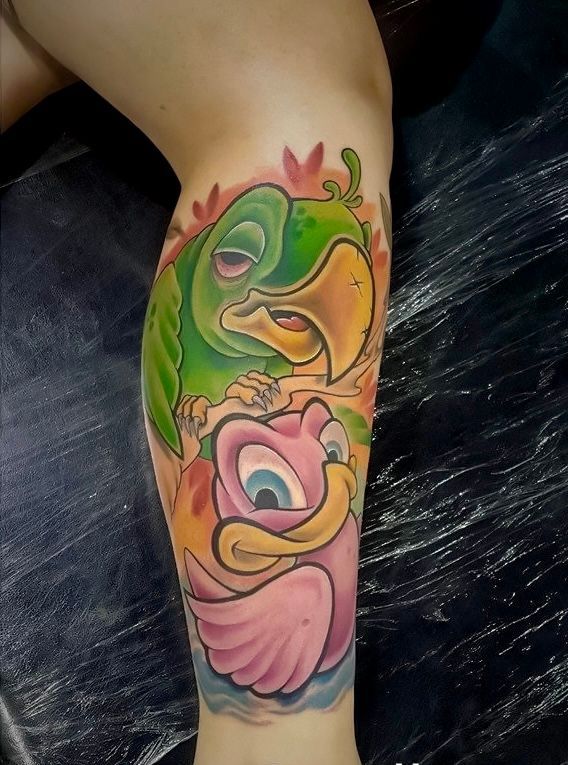 A vibrant tattoo on a leg featuring a sleepy green parrot perched above a wide-eyed, smiling pink bird.