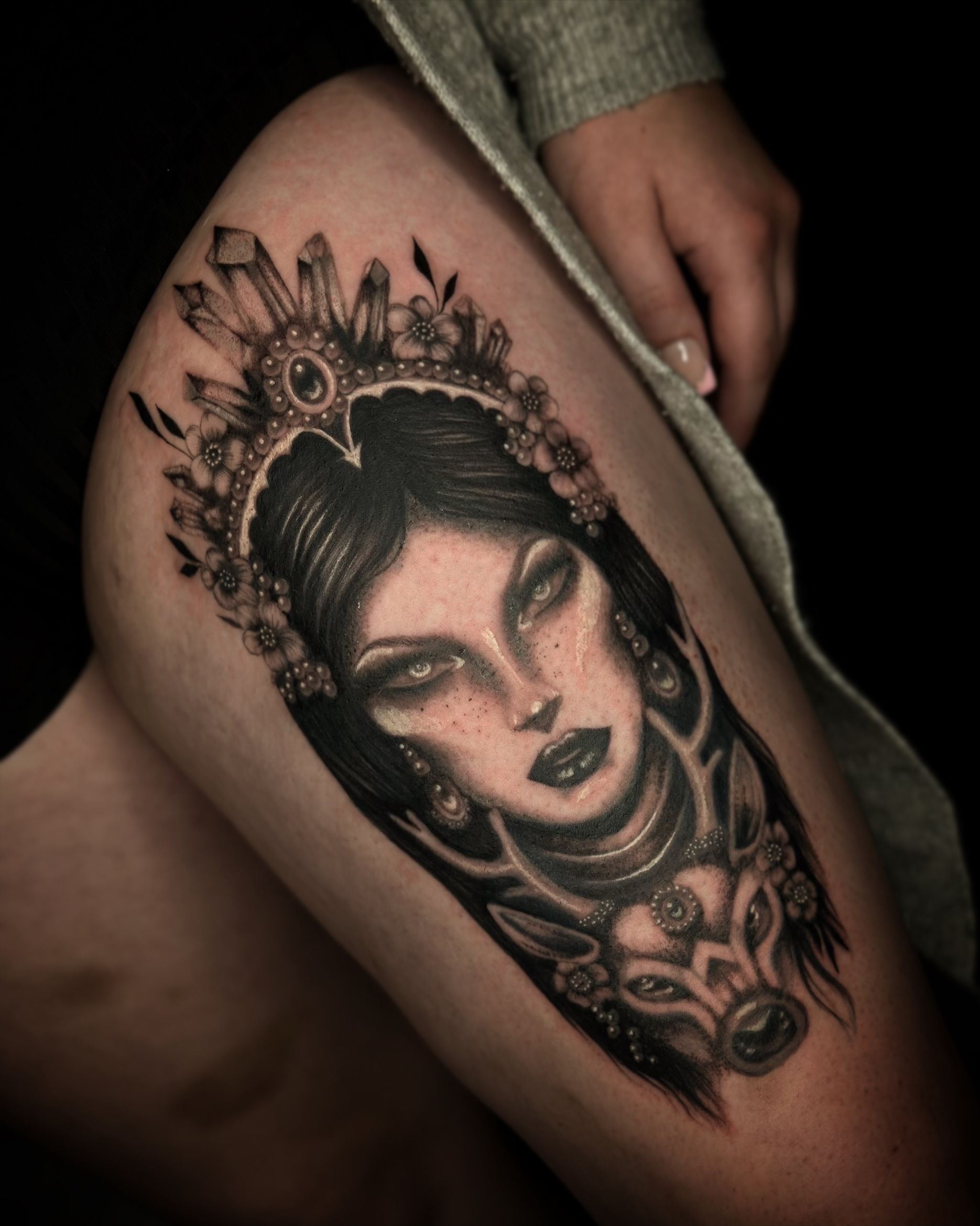 Black and grey ink tattoo on a thigh featuring a woman’s face wearing a crystal-adorned crown above a deer-like animal.