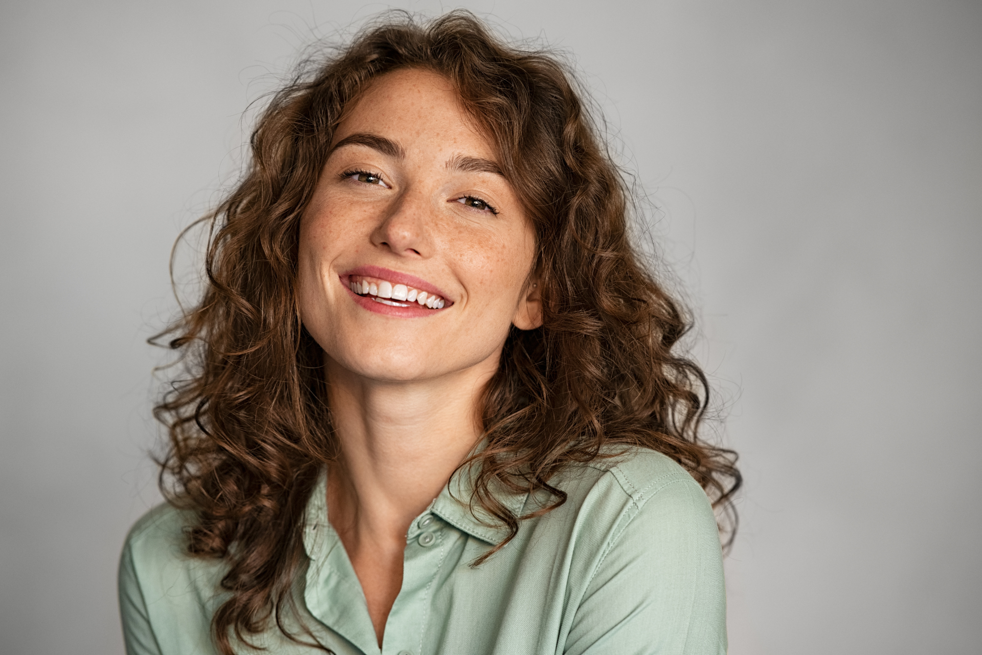 Smiling person with curly brown hair in a light green shirt against a gray background