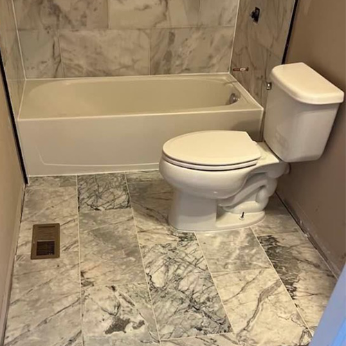 A bathroom with a white bathtub and toilet, featuring gray and white marble-style flooring and wall tiling.
