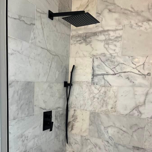 A matte black shower head and handheld sprayer mounted on a wall with white and grey marbled tile.