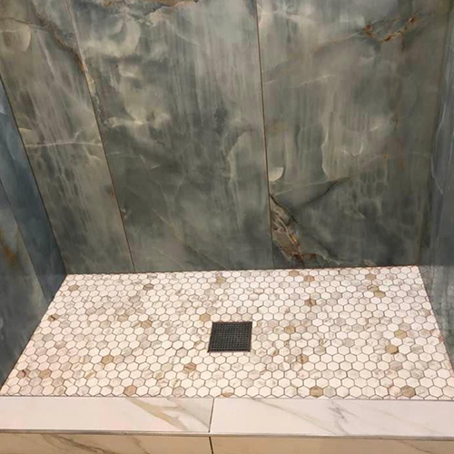 A shower floor with white and tan penny tiles, a centered square drain, and blue-gray marble-look wall tiles.