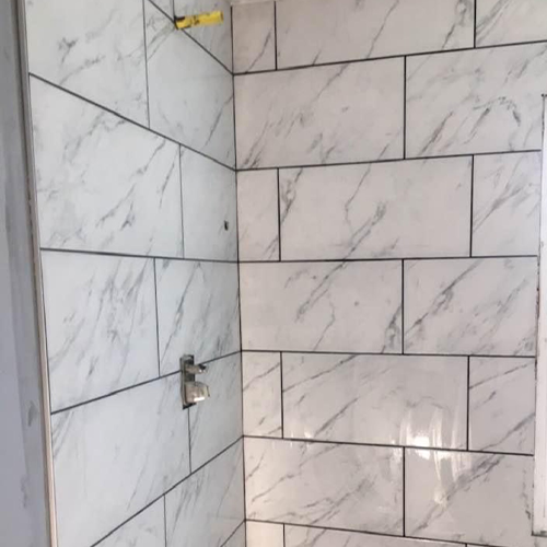A corner of a shower featuring large white tiles with grey marble-like veining, installed in a running bond pattern.