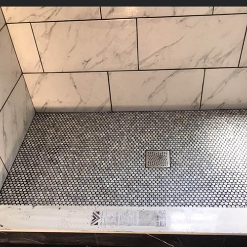 A shower stall with large white marble-patterned wall tiles and a small dark penny-round tile floor with a metal drain.