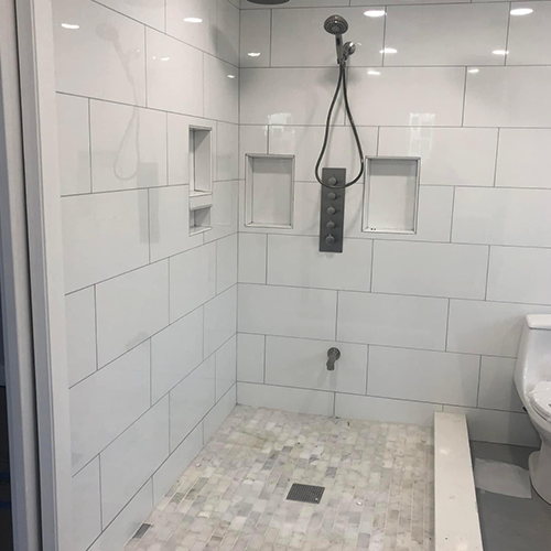 A walk-in shower with white tiled walls, three recessed storage niches, a handheld showerhead, and a stone mosaic floor.