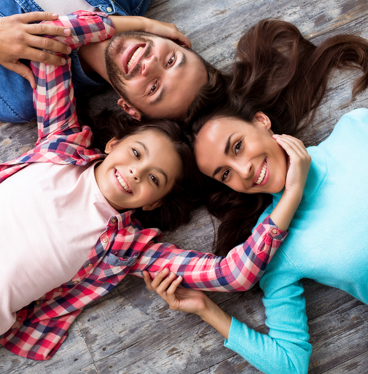 Three smiling friends lying together on a wooden floor, looking up at the camera.