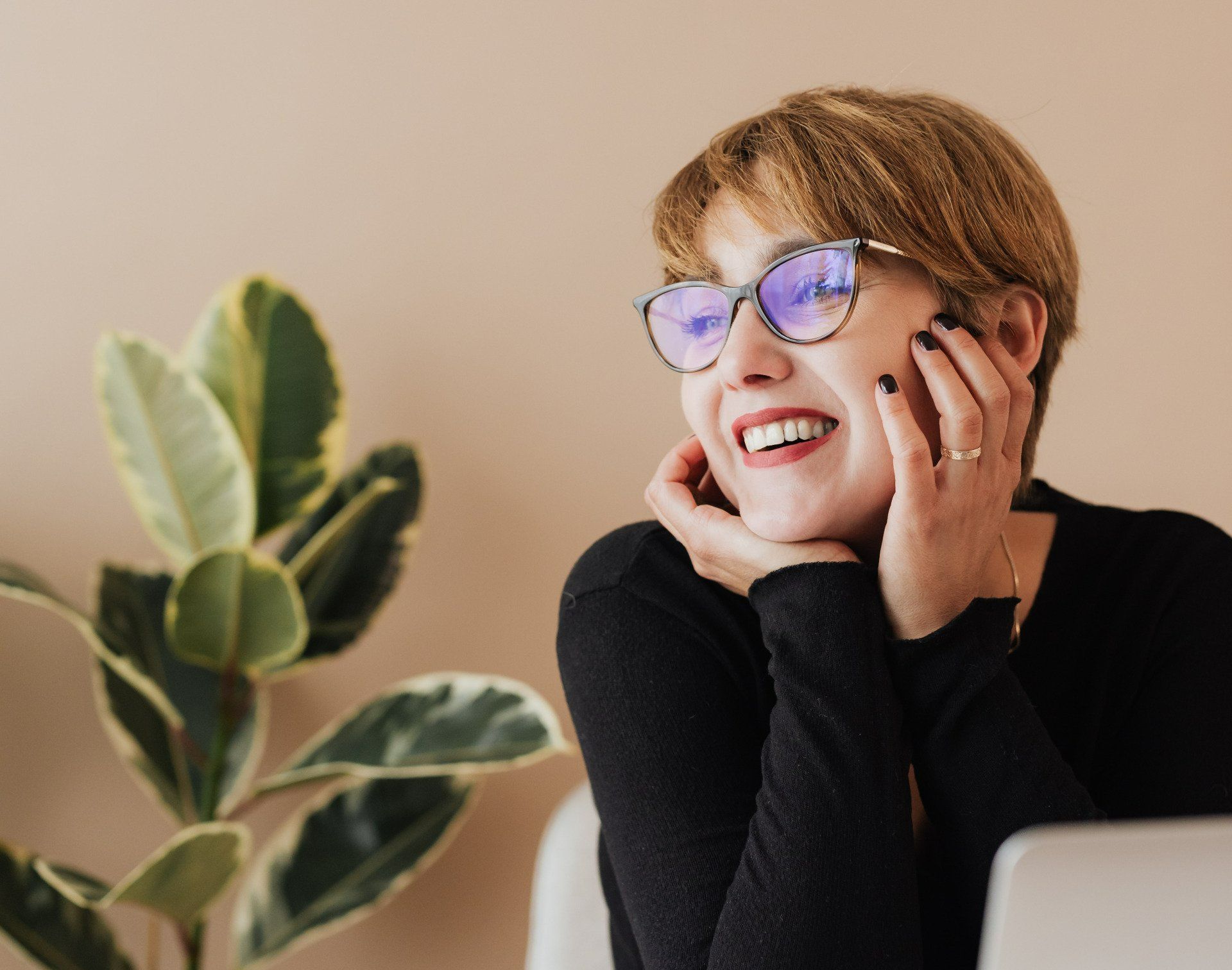 Smiling person with glasses resting their face on their hands beside a leafy plant