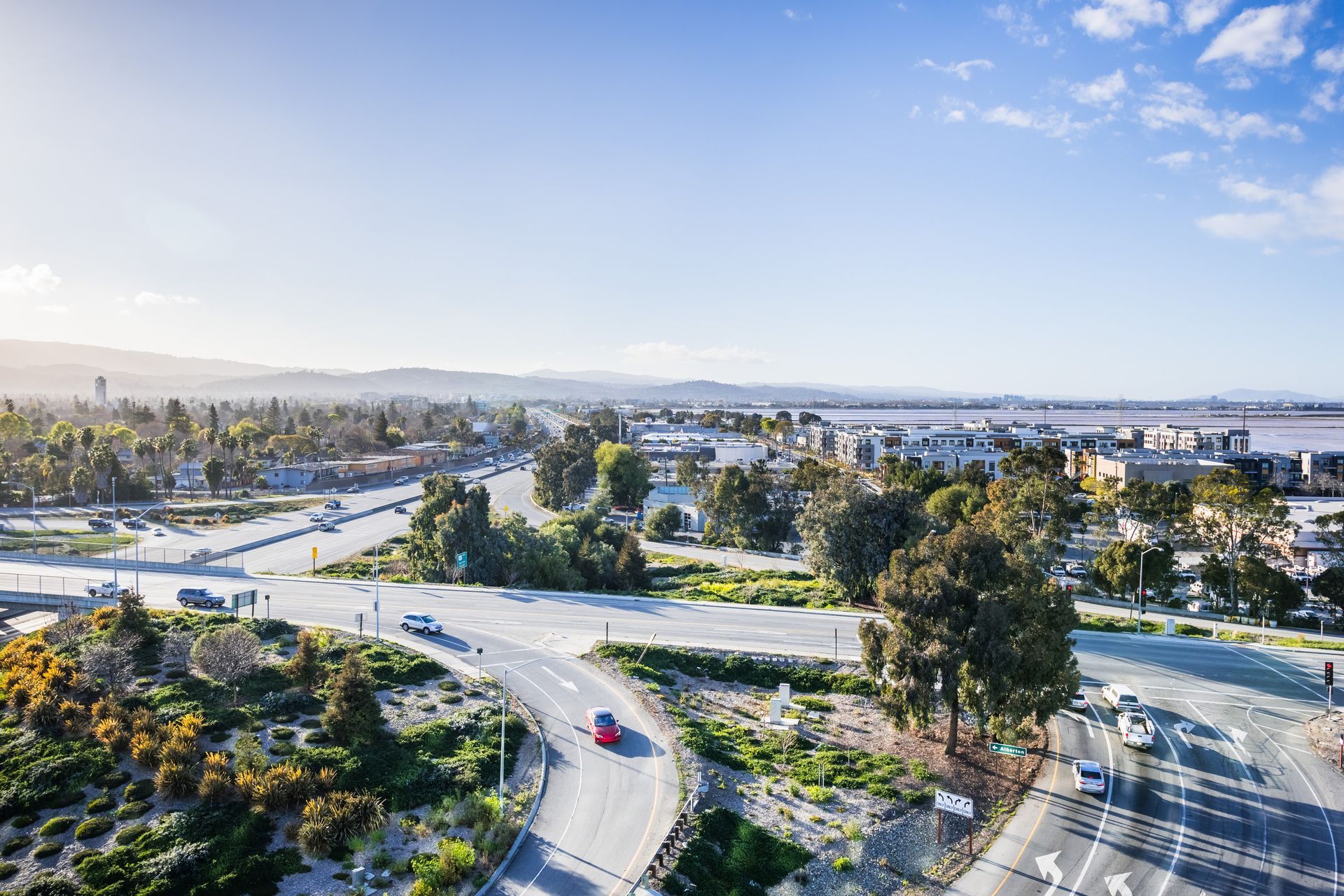 Wide aerial view of a winding road through a sunny green urban landscape with distant city buildings
