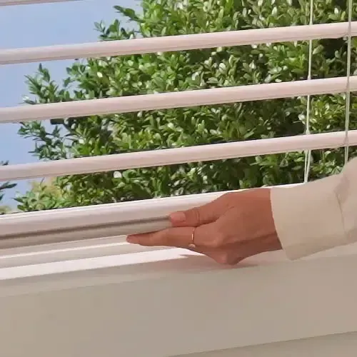 Person's hands adjusting white horizontal blinds, sunlight and green foliage visible through the slats.