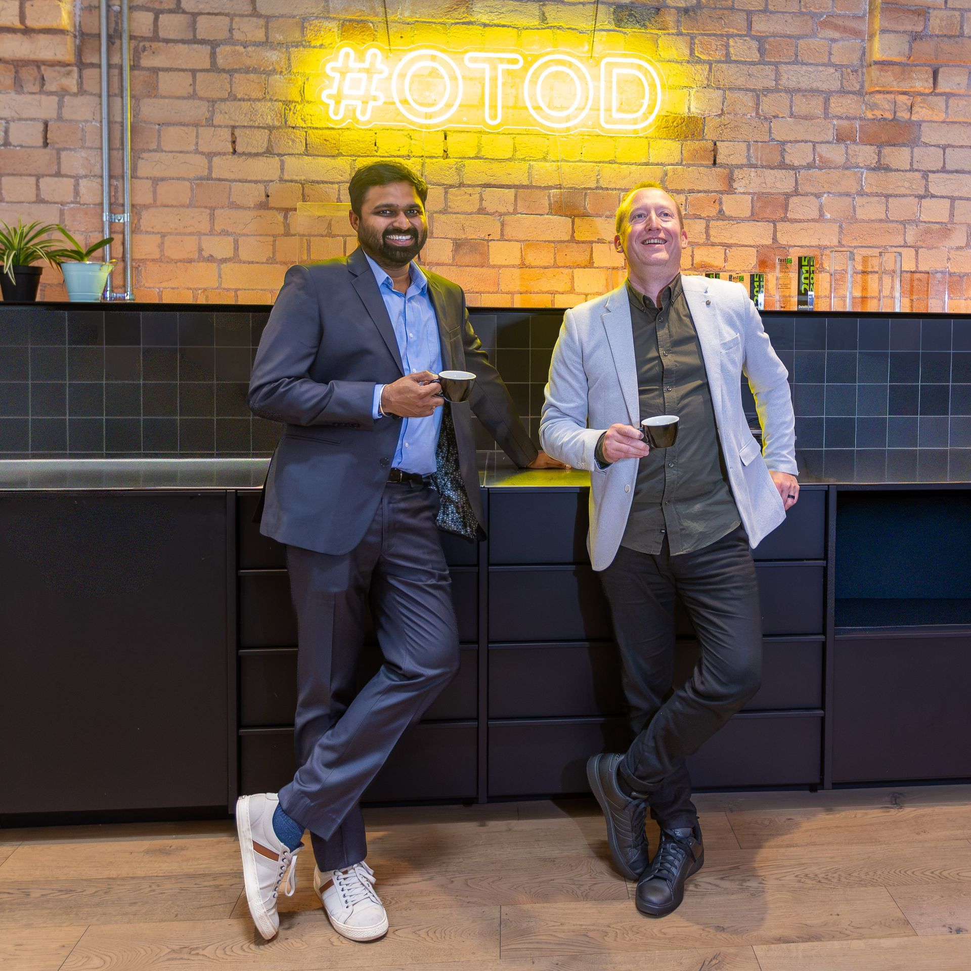 Two men in suits leaning against a black counter beneath a yellow neon “HOTDOG” sign.