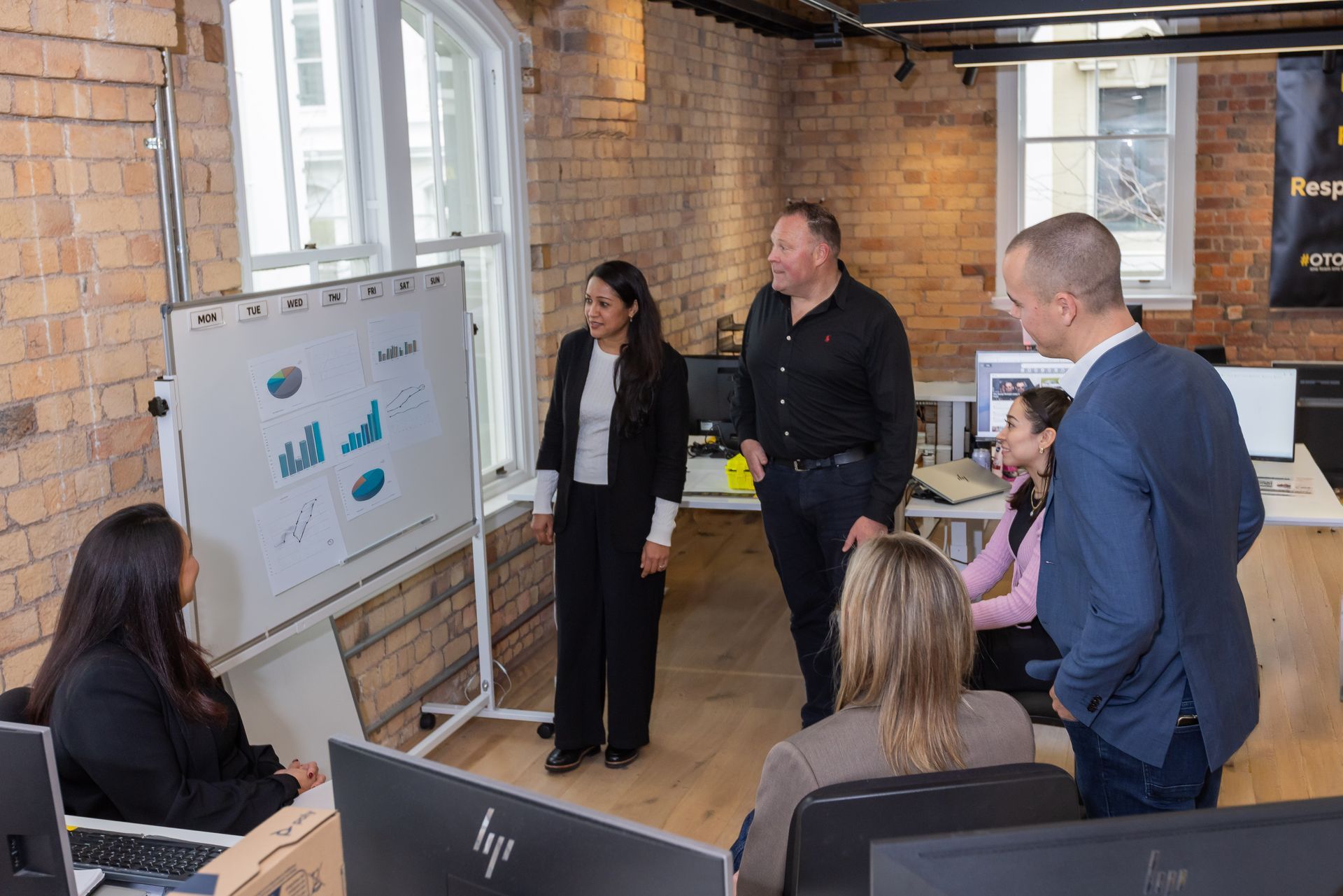 Five people seated at a table with laptops and drinks in a brick-walled meeting room.