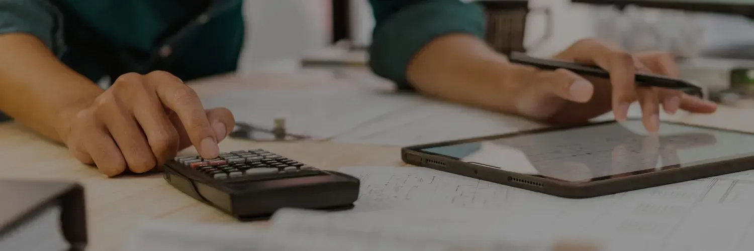 People writing on paper at a table with calculators, one using a pen near a notebook.