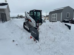 Skid steer with snowplow clearing a snow-covered driveway between houses