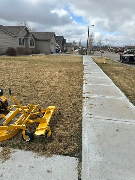 Sidewalk beside a grassy residential lot, with a yellow lawn mower in the foreground and cloudy sky overhead