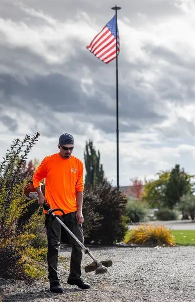 Person in orange shirt using a string trimmer on gravel beneath an American flag under cloudy sky