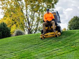 Person mowing a green hillside lawn on a yellow riding mower, wearing an orange shirt and blue cap.
