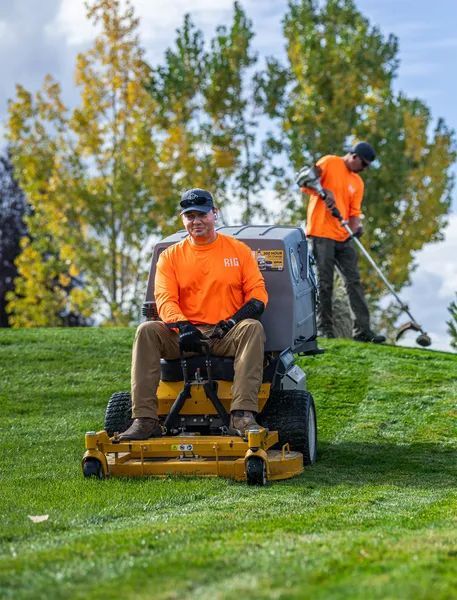 Two landscapers in orange shirts mow and trim a grassy slope with autumn trees behind them.