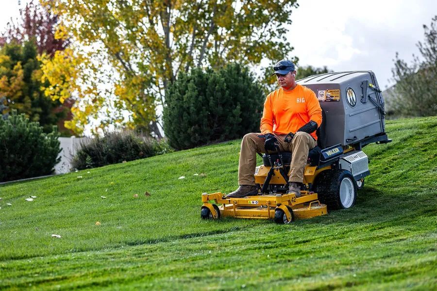 Worker in orange shirt mowing a grassy slope with a riding lawn mower.