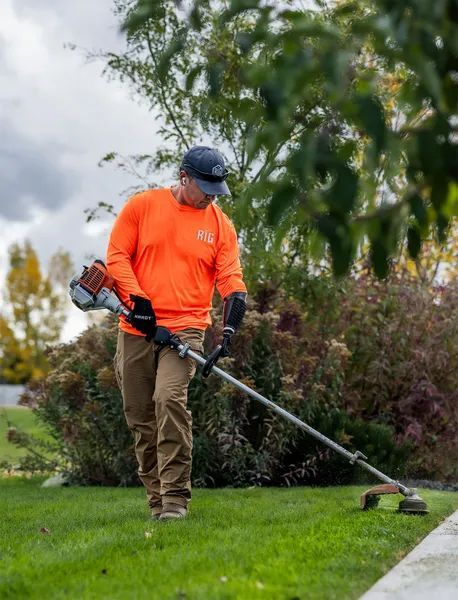 Worker in orange shirt trimming grass along a sidewalk with a string trimmer.