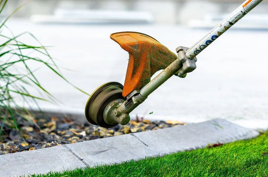 Orange string trimmer cutting grass along a curb beside a sidewalk and gravel edge