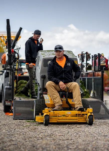 Two people on yellow lawn mowers in a gravel lot, with one seated in front and another standing behind.