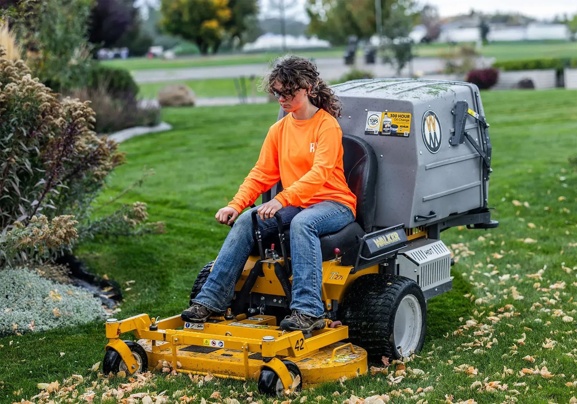 Person in orange shirt mowing a grassy lawn with a ride-on mower in a park-like setting