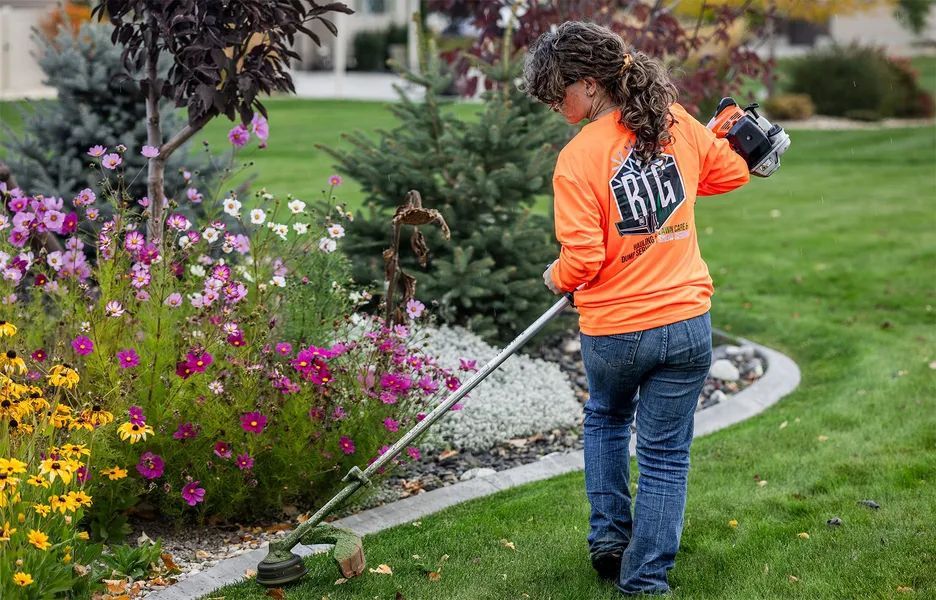 Person in orange shirt using a string trimmer beside a flower bed in a garden