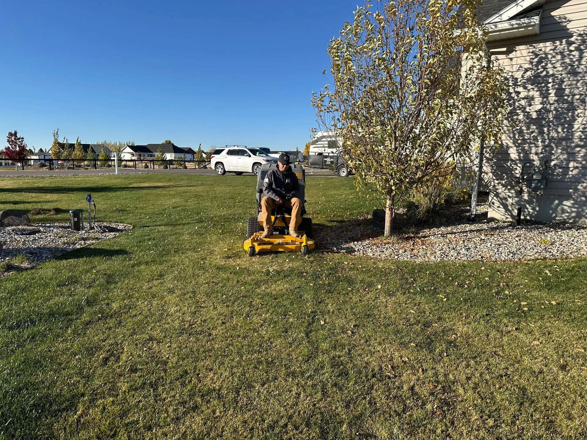 Person mowing a grassy yard with a yellow mower beside a house and tree