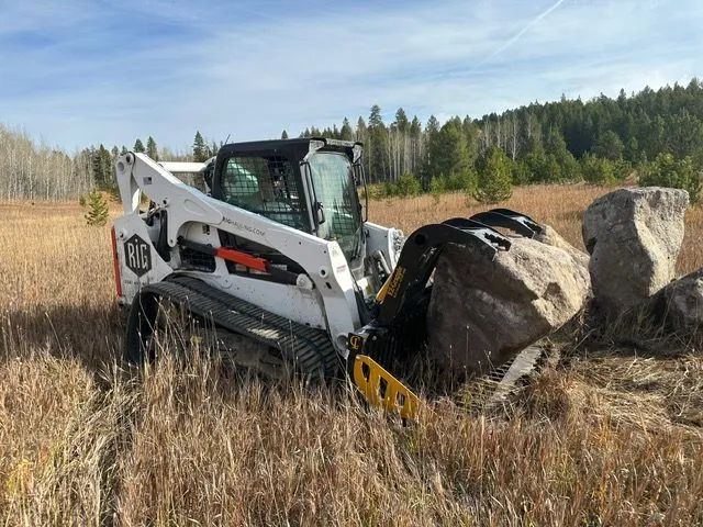 Skid steer loader pushing a large boulder in a grassy field with trees in the background