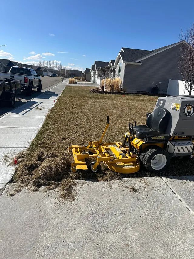 Yellow lawn mower on a driveway beside a brown yard in a suburban neighborhood under a blue sky
