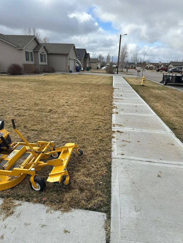 Wet suburban sidewalk beside a house, with a yellow lawn mower on the grass under cloudy skies.