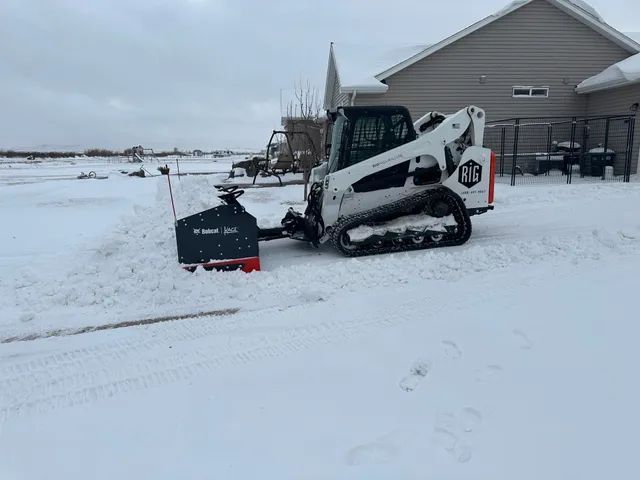 White skid steer with snow blade clearing snow beside a house in a snowy yard